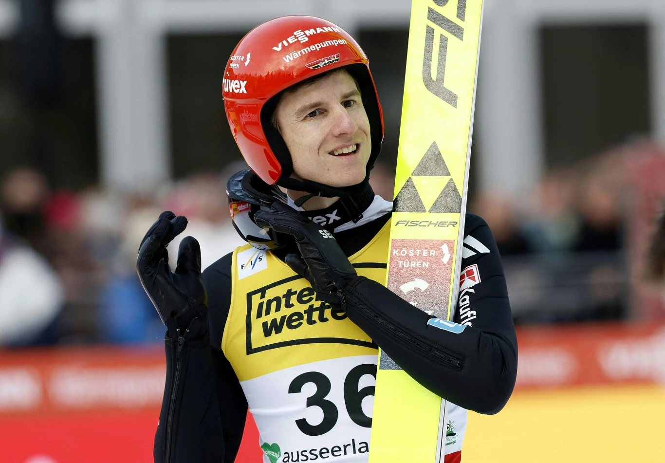 skoczek narciarski w czerwonym kasku i czarnym kombinezonie z numerem startowym 36 trzyma narty na ramieniu, uśmiecha się i wykonuje gest dłonią, w tle rozmyty stadion