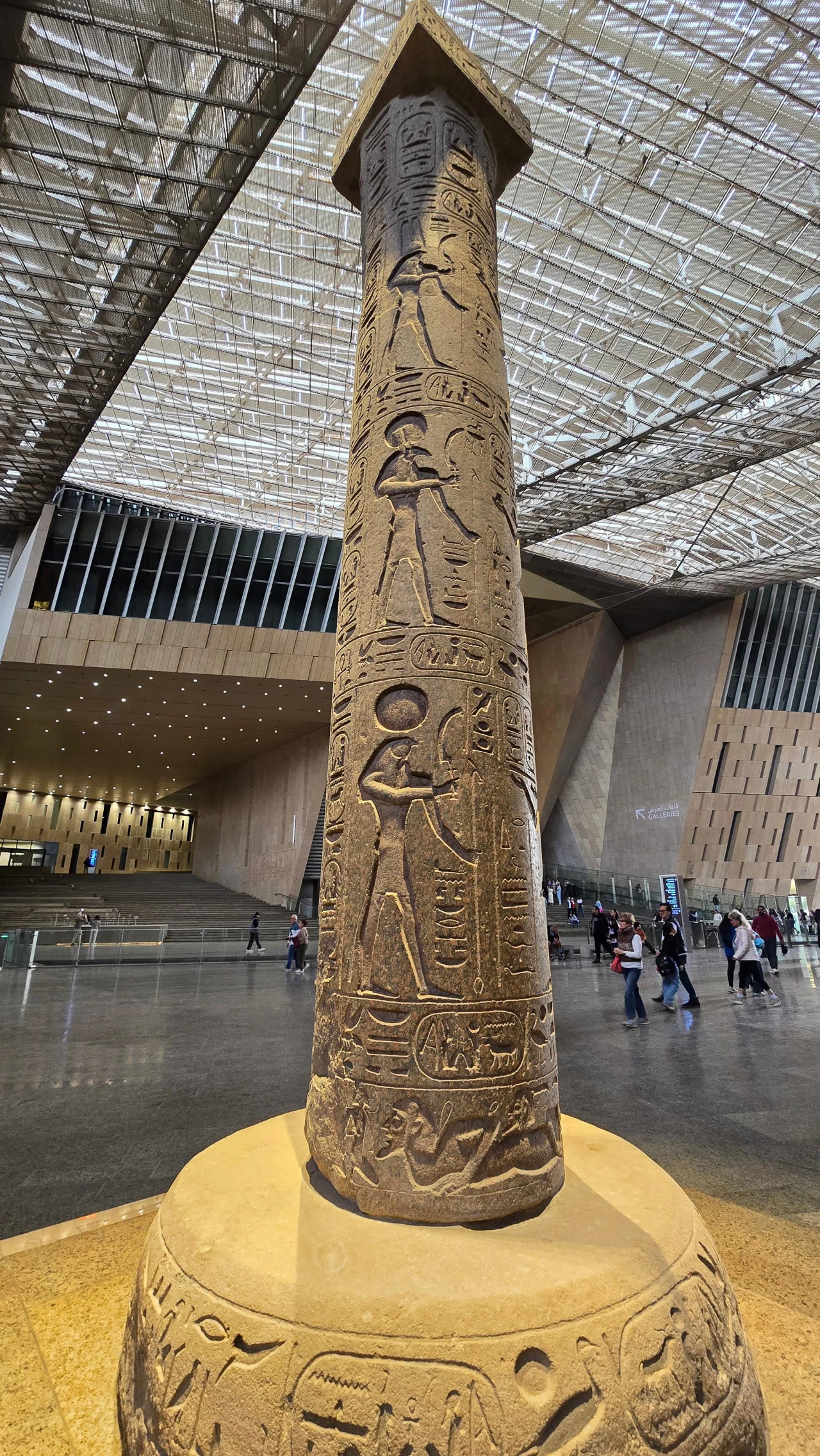 wysoki kamienny obelisk z wyrytymi egipskimi hieroglifami i postaciami bóstw, ustawiony w nowoczesnym przestronnym wnętrzu muzeum z przeszklonym dachem, wokół którego przechodzą zwiedzający