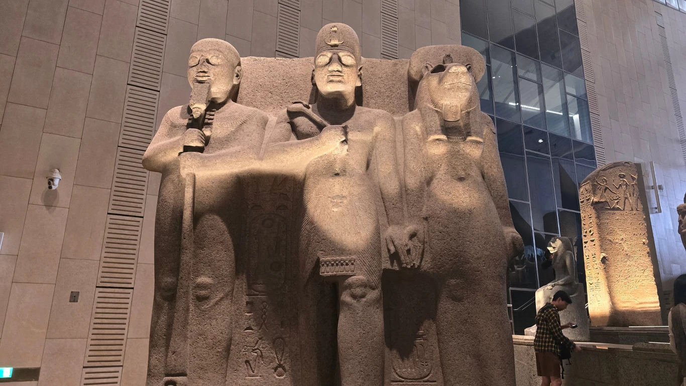 trzy masywne starożytne egipskie posągi z piaskowca stojące obok siebie, każdy z rękami skrzyżowanymi na piersi, wykonane z dużą dbałością o detale, ustawione w nowoczesnym wnętrzu muzealnym pełnym światła oraz przejrzystych powierzchni