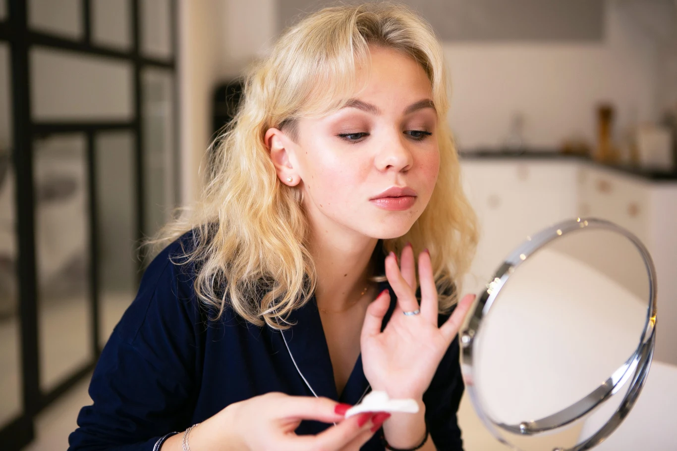 Młoda kobieta o blond włosach siedzi przy stoliku i patrzy w lusterko, w jednej ręce trzyma wacik kosmetyczny, a drugą delikatnie dotyka twarzy; jej twarz jest skupiona na czynności pielęgnacyjnej lub makijażowej.