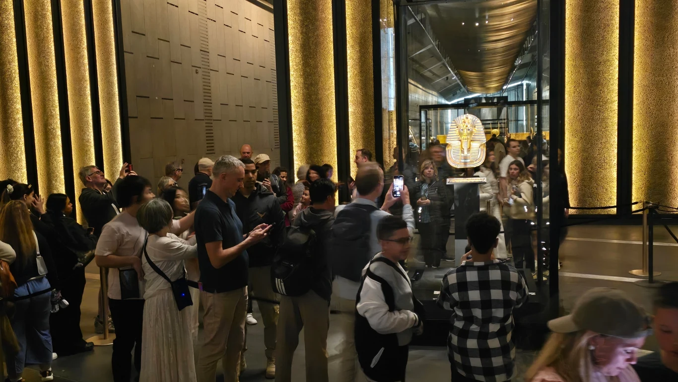 tłum ludzi zgromadzony wokół szklanej gabloty, w której eksponowana jest złota maska Faraona Toutanchamona, liczne osoby robią zdjęcia lub nagrywają wideo, tło stanowią nowoczesne, podświetlone ściany muzealne