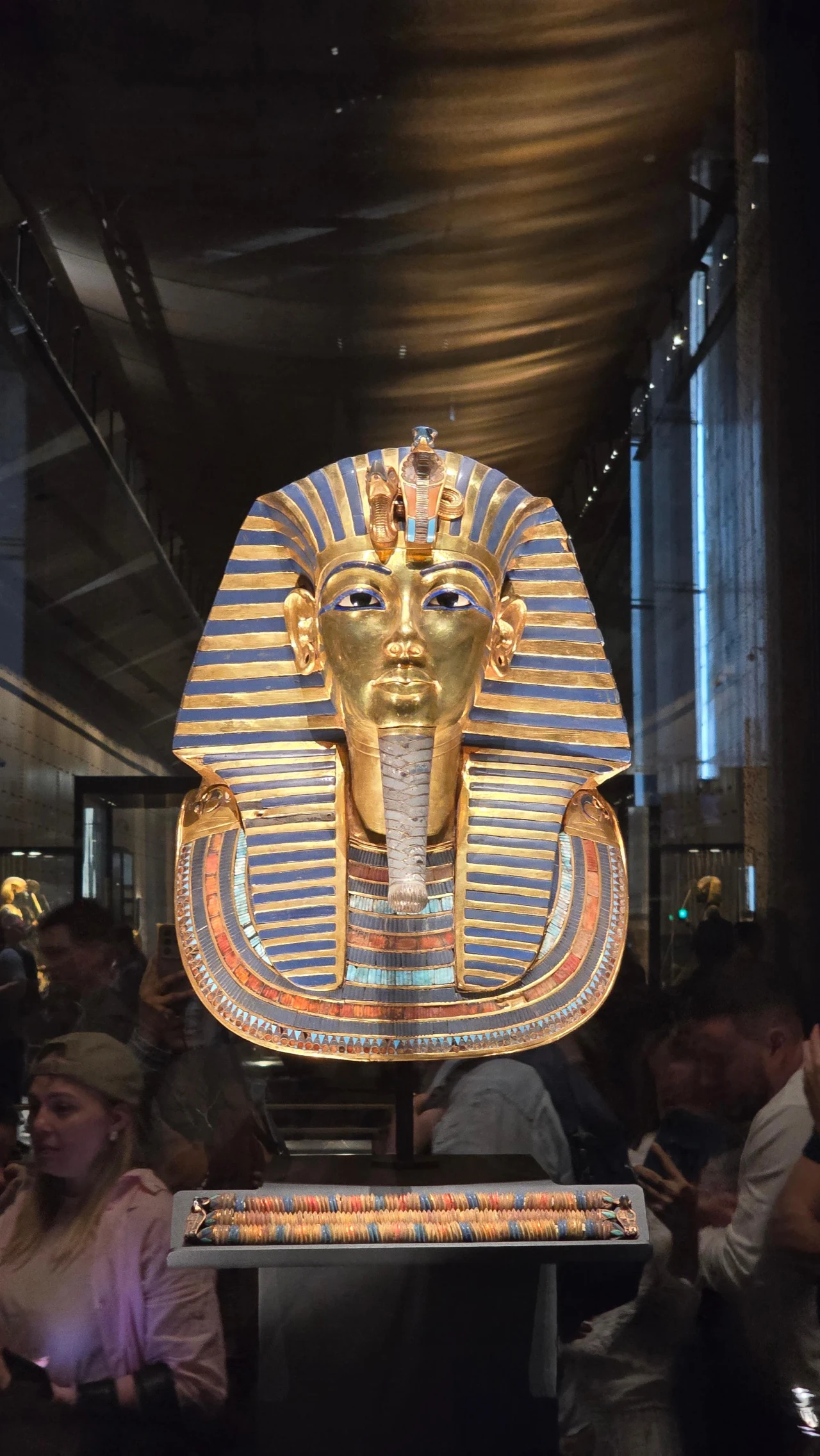 Złota maska Tutanchamona ozdobiona kolorowymi kamieniami szlachetnymi i niebieskimi paskami, otoczona przez zwiedzających wewnątrz muzeum.