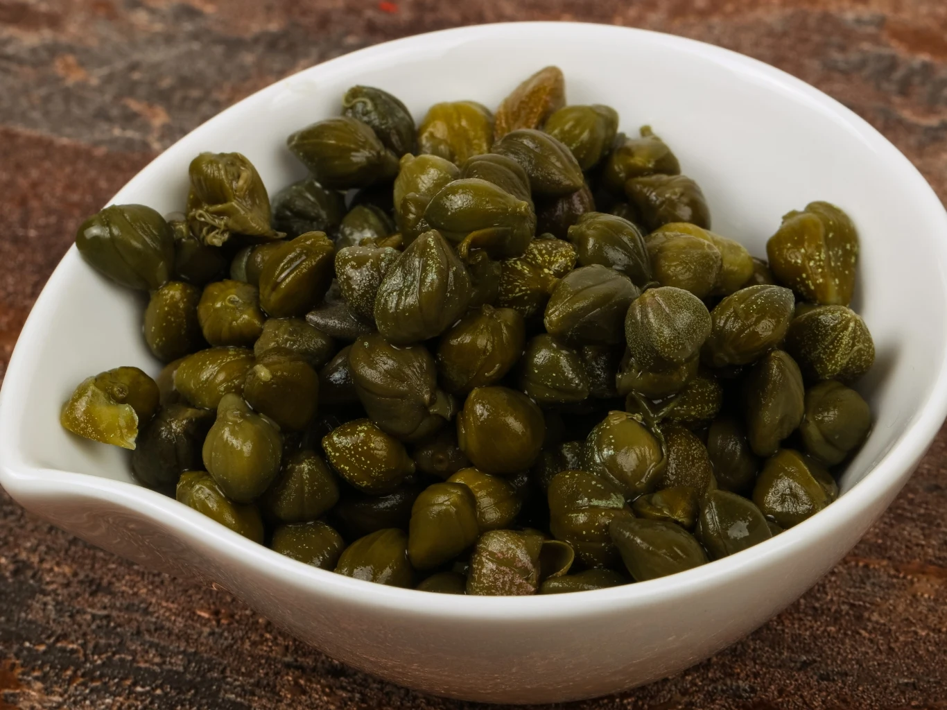 Jedz jako przekąskę lub dodaj do sałatki, gdy masz wysoki cukier i cholesterol. Kapary w słonej zalewie w białej miseczce na ciemnym tle, do sałatek i jako przekąska dla osób z wysokim cukrem i cholesterolem.