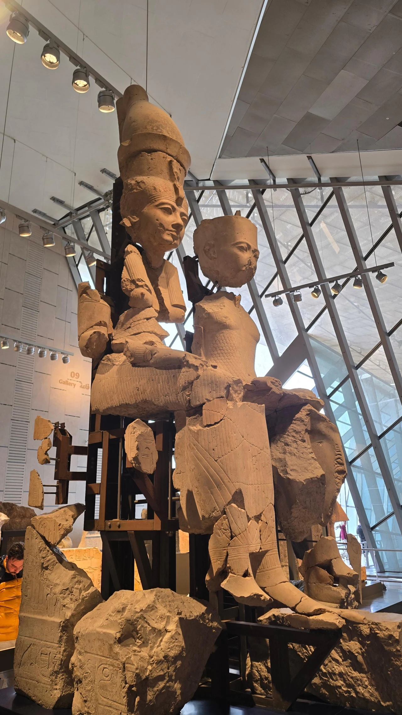 Dwie monumentalne kamienne rzeźby siedzących postaci ze starożytnego Egiptu, prezentowane na stalowej konstrukcji wspierającej, z widocznymi uszkodzeniami i brakującymi fragmentami, ustawione w nowoczesnym wnętrzu muzeum z przeszkloną ścianą w tle.