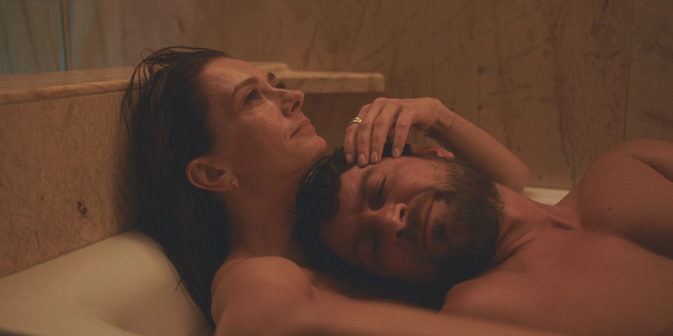 Kobieta obejmuje leżącego na jej klatce piersiowej mężczyznę, oboje zanurzeni w wannie, w ciepłej, intymnej atmosferze; na dłoni kobiety widoczny pierścionek.