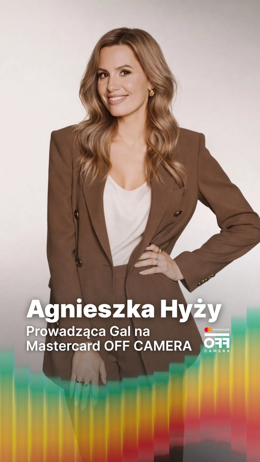 Kobieta z długimi blond włosami ubrana w elegancki, brązowy garnitur z jasną bluzką, stojąca na tle jasnego, jednolitego tła, z napisem informującym o jej roli jako prowadzącej galę Mastercard OFF CAMERA.