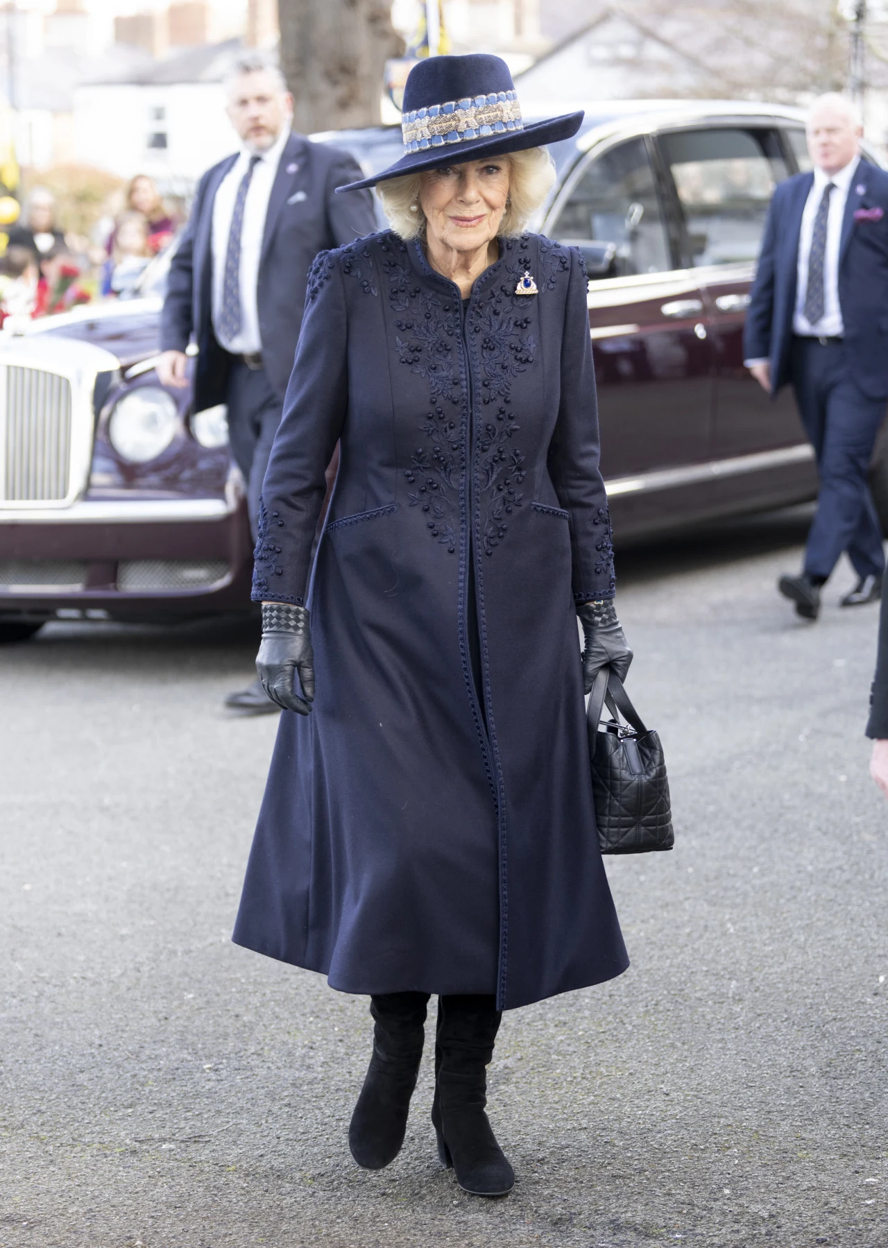 Elegancka kobieta w granatowym płaszczu ozdobionym haftami, kapeluszu z szerokim rondem oraz czarnych rękawiczkach i butach stoi na ulicy, trzymając czarną torebkę; w tle widać luksusowy samochód oraz kilku mężczyzn w garniturach.