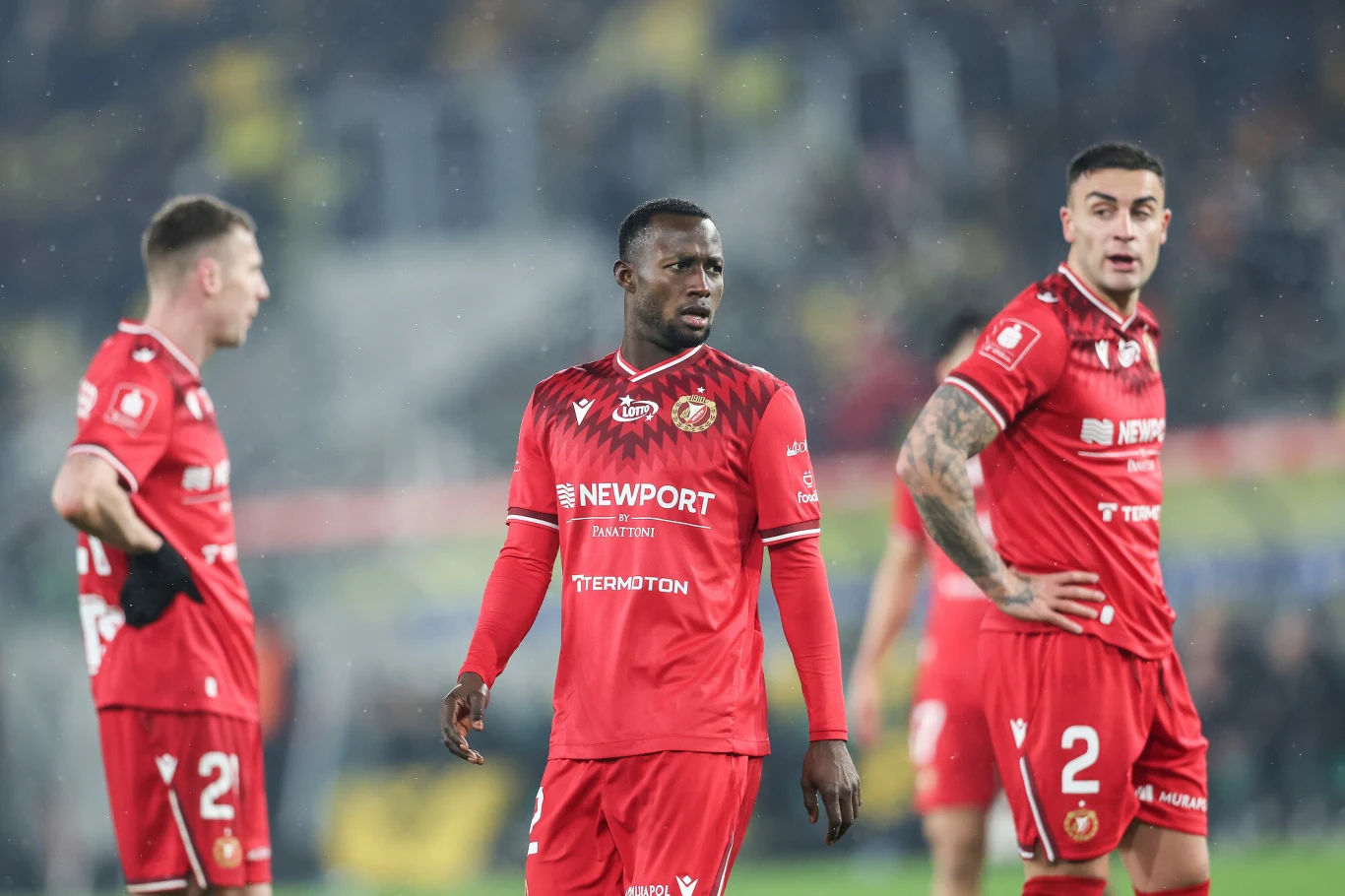 trzech piłkarzy w czerwonych strojach drużyny Widzew Łódź stoi na murawie podczas meczu, na twarzach widać skupienie i napięcie, w tle rozmazane sylwetki kibiców na trybunach