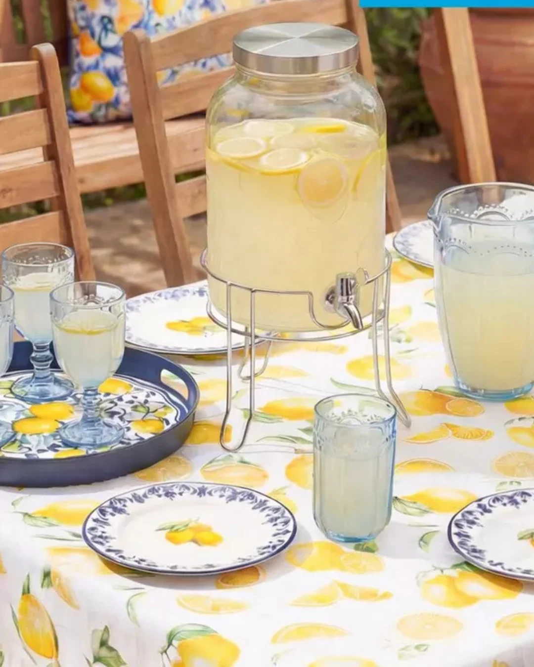 Duży szklany dzban z lemoniadą i plasterkami cytryny ustawiony na metalowym stojaku pośrodku stołu, obok szklanki z napojem, dzbanek oraz talerze z motywem cytryn na obrzeżach i tkanina stołowa ozdobiona cytrynami, otoczone drewnianymi krzesłami.