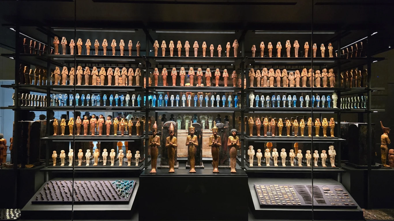 Gabloty muzealne wypełnione licznymi figurkami przedstawiającymi postacie z czasów starożytnego Egiptu, ułożone w równych rzędach, z przodu znajdują się wieksze figury oraz eksponaty biżuterii i drobnych przedmiotów użytkowych.