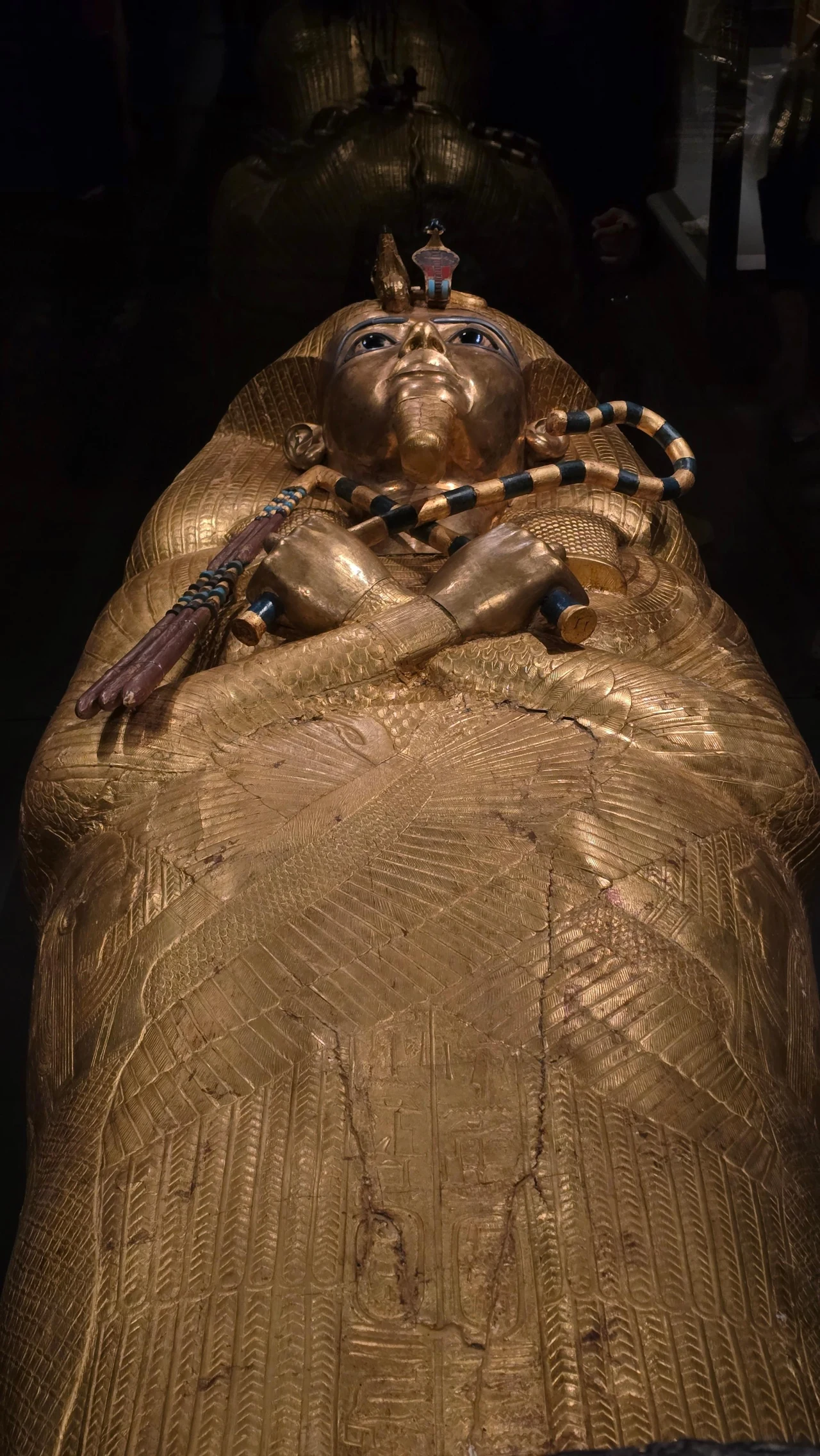 Bogato zdobiona złota trumna w kształcie ludzkiej postaci z charakterystyczną maską na twarzy, skrzyżowanymi na piersi ramionami oraz insygniami egipskiego faraona, otoczona ciemnym tłem.