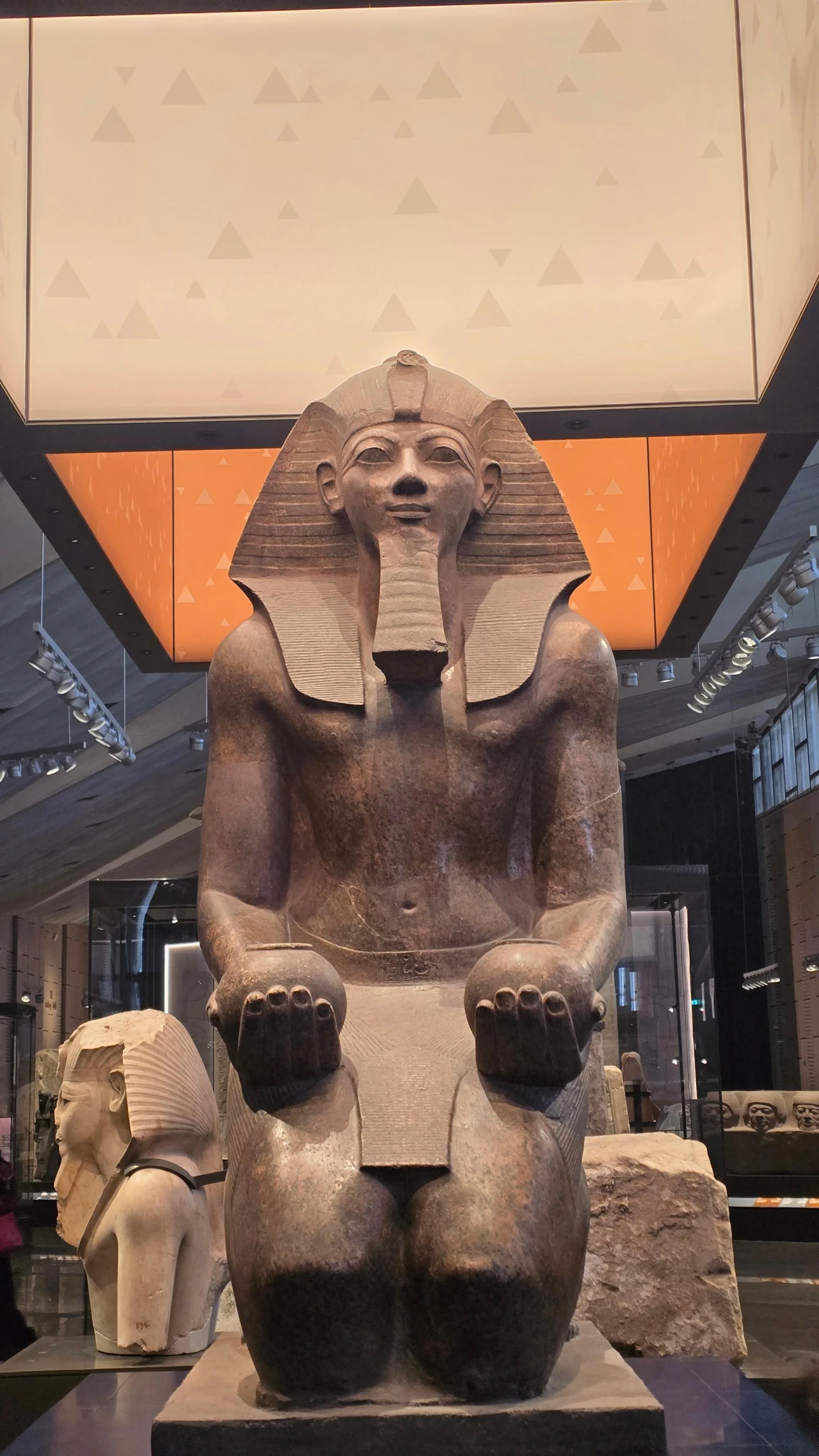 Monumentalna rzeźba egipskiego faraona w pozycji klęczącej, z dłońmi opartymi na kolanach i charakterystyczną królewską nakryciem głowy, ustawiona w muzealnej sali z nowoczesnym oświetleniem w tle.