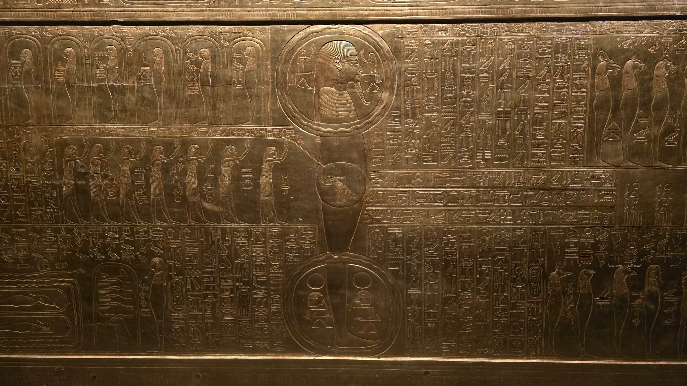 starożytna egipska płyta pokryta szczegółowymi hieroglifami oraz reliefami, w tym postaci siedzącej osoby z nimbem wokół głowy i licznymi postaciami stojącymi wokół, uzupełniona symbolami i postaciami ptaków, całość wykonana z metalu o złotym połysku
