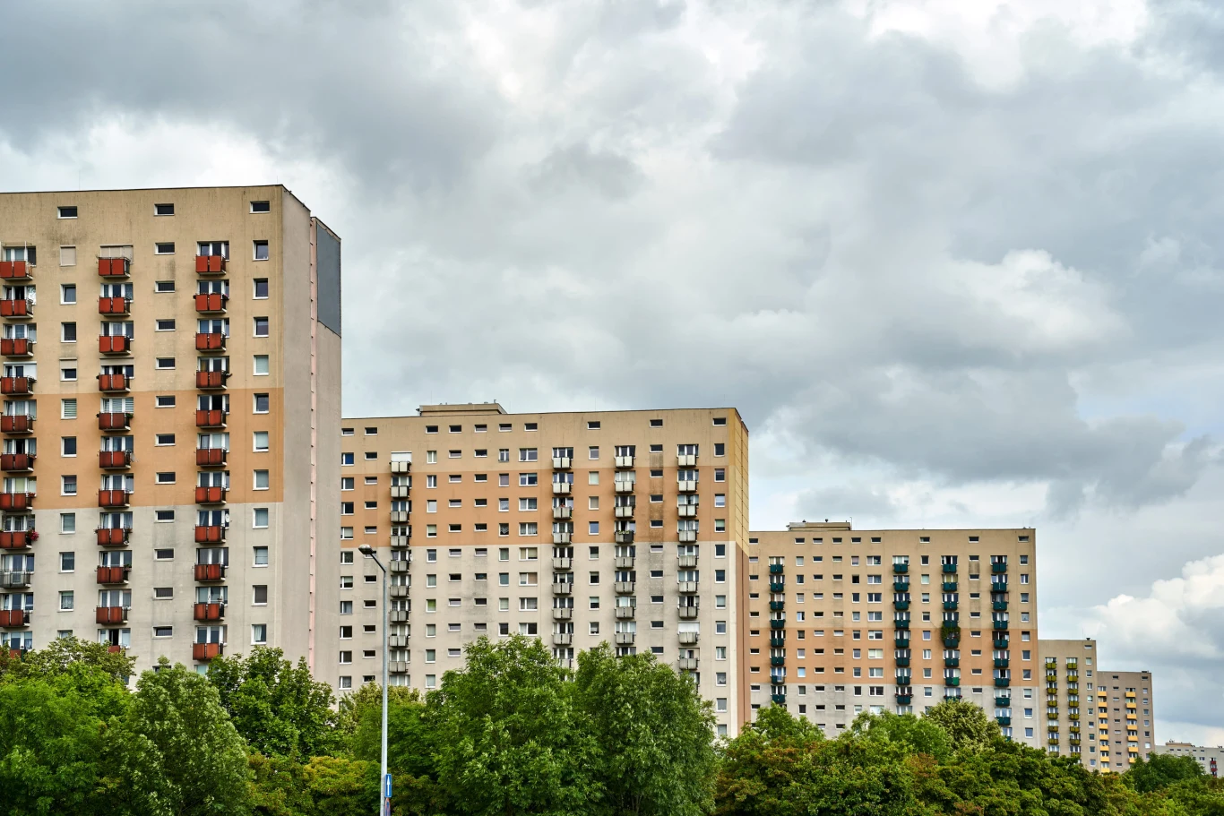 Wysokie bloki mieszkalne o jasnej elewacji z rzędami okien i balkonów, otoczone gęstą zielenią drzew na pierwszym planie, pod pochmurnym niebem.