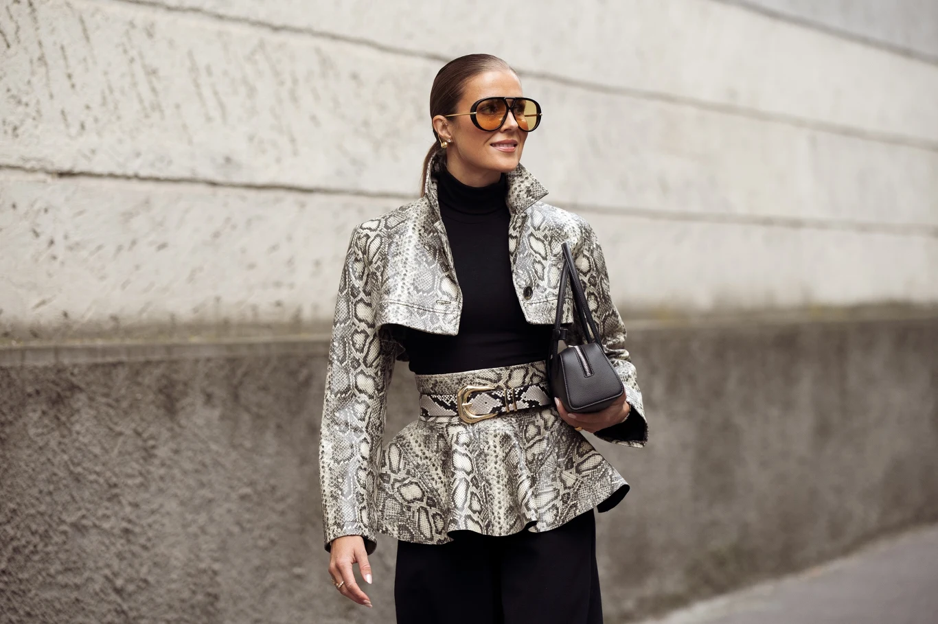 kobieta w okularach przeciwsłonecznych ubrana w stylową kurtkę o wzorze skóry węża, czarny golf i spodnie, trzymająca elegancką torebkę na ramieniu, idzie wzdłuż jasnej, betonowej ściany