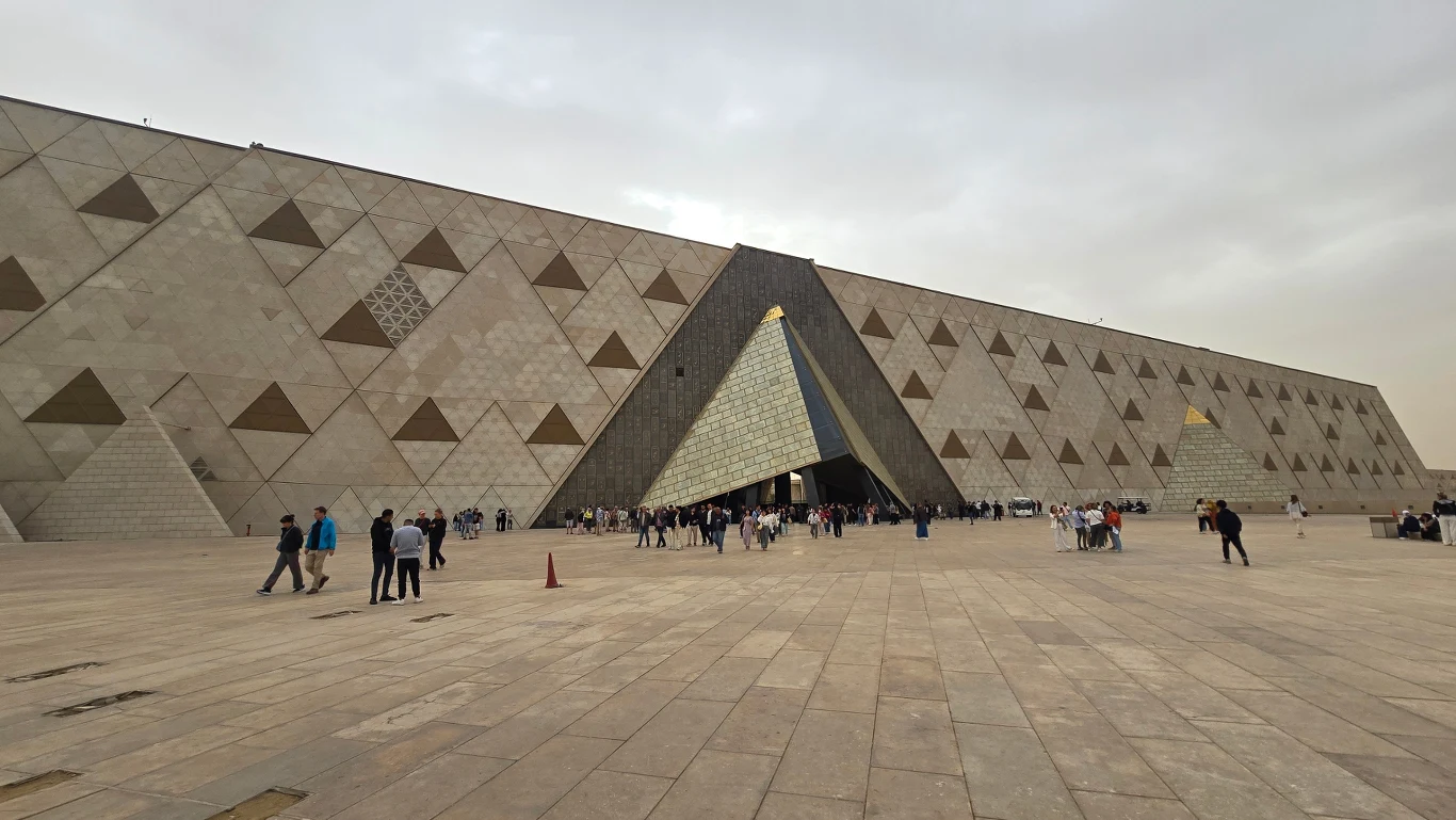 Monumentalny budynek o geometrycznych zdobieniach na elewacji, z centralnym, trójkątnym wejściem, przed którym gromadzą się ludzie na rozległym, wyłożonym płytami placu. Wielkie Muzeum Egipskie.