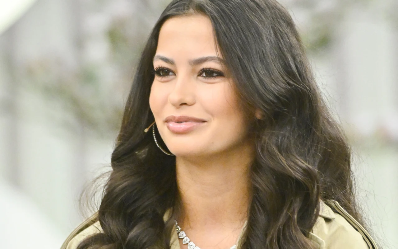 kobieta z długimi, ciemnymi, falowanymi włosami, delikatnym makijażem i łagodnym uśmiechem, ubrana w beżową kurtkę oraz naszyjnik z błyszczącymi kamieniami, z mikrofonem przy twarzy