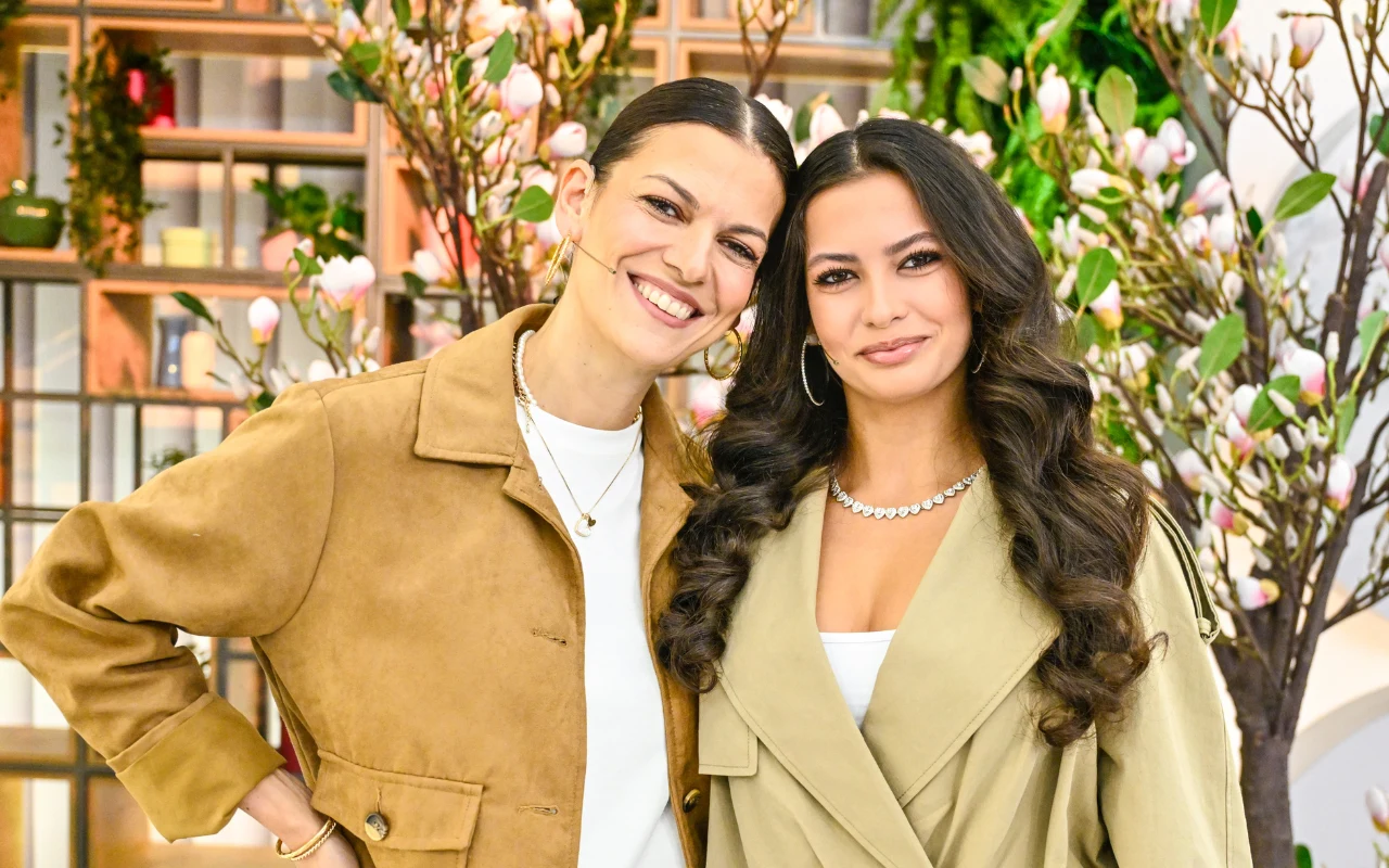 dwie uśmiechnięte kobiety stoją obok siebie w jasnych, stylowych płaszczach, otoczone wiosenną dekoracją z kwitnących gałęzi i zieleni