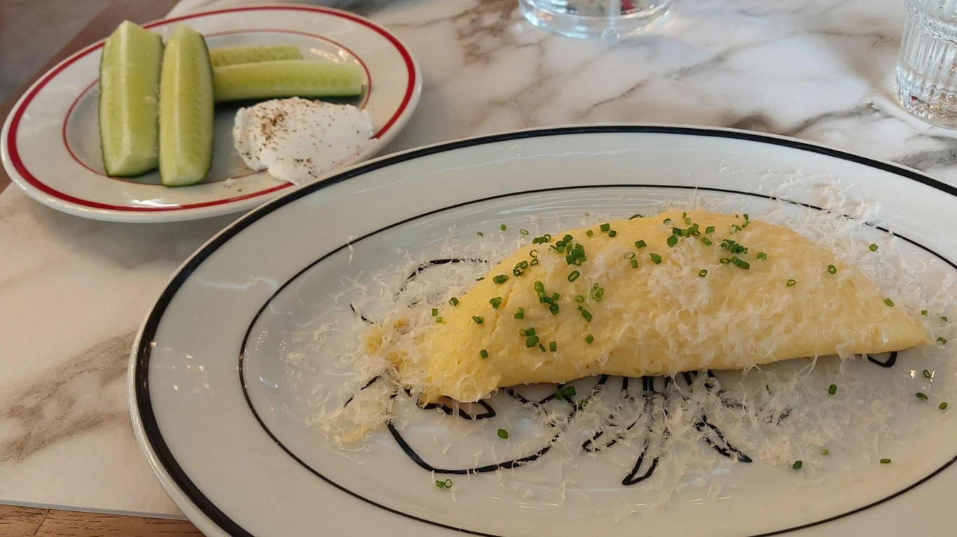 puszysty omlet posypany świeżo startym parmezanem i szczypiorkiem na białym talerzu, ozdobiony cienkimi liniami sosu balsamicznego, obok na małym talerzyku plastry zielonego ogórka z porcją białego serka i pieprzem