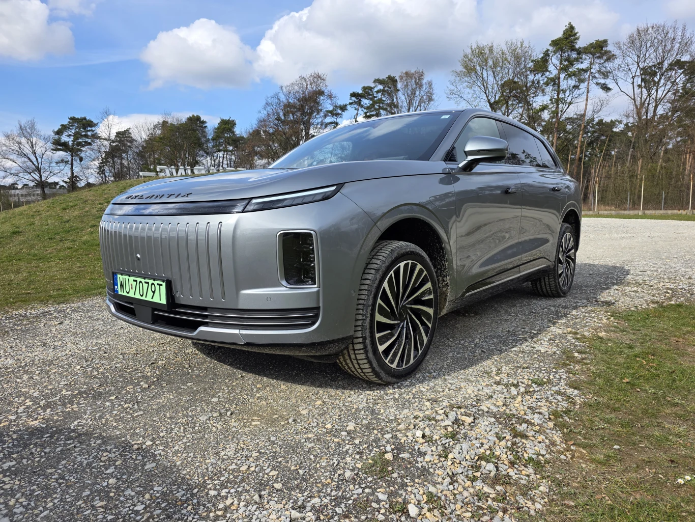 srebrny, nowoczesny SUV z charakterystycznym, pionowym grillem i wąskimi, poziomymi światłami LED, stoi na żwirowym placu w otoczeniu niskiego lasku oraz nieba z rozproszonymi chmurami