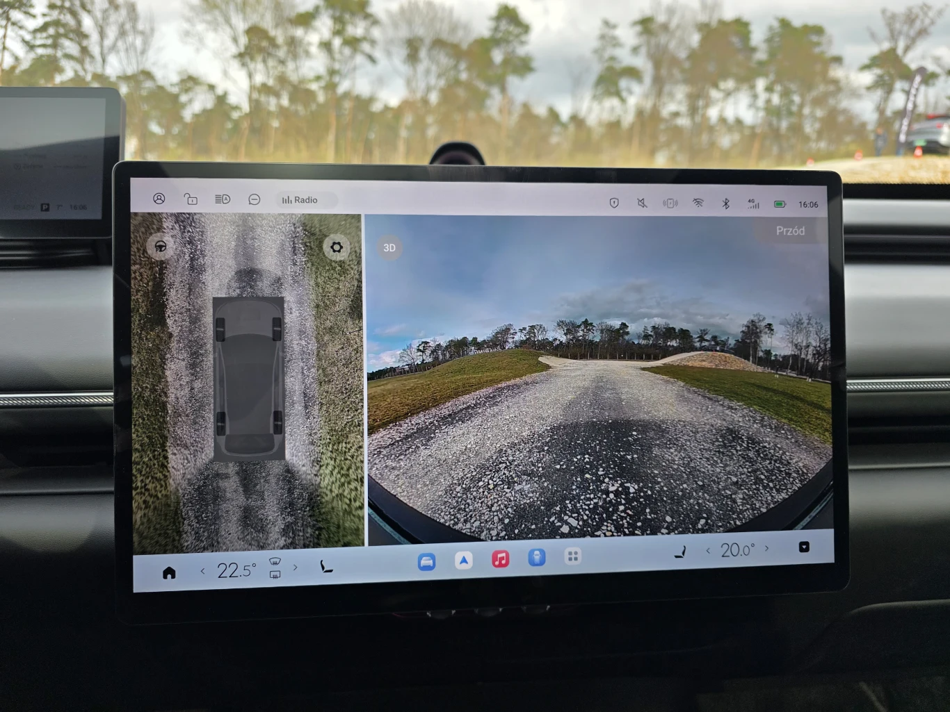 duży ekran dotykowy w samochodzie wyświetlający system kamer 360 stopni, pokazujący widok z lotu ptaka na auto oraz szeroki obraz drogi przed pojazdem, otoczonej drzewami i trawiastymi poboczami
