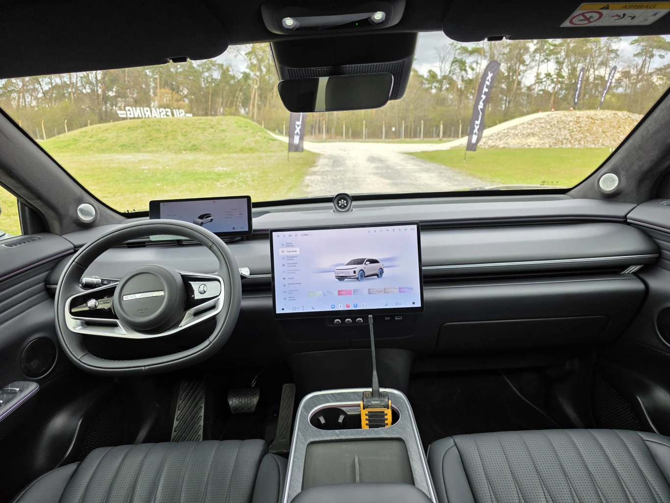 nowoczesne wnętrze samochodu elektrycznego z dużym centralnym ekranem dotykowym, cyfrowym wyświetlaczem za kierownicą i minimalistyczną deską rozdzielczą; przez przednią szybę widoczna jest leśna droga oraz zieleń wokół