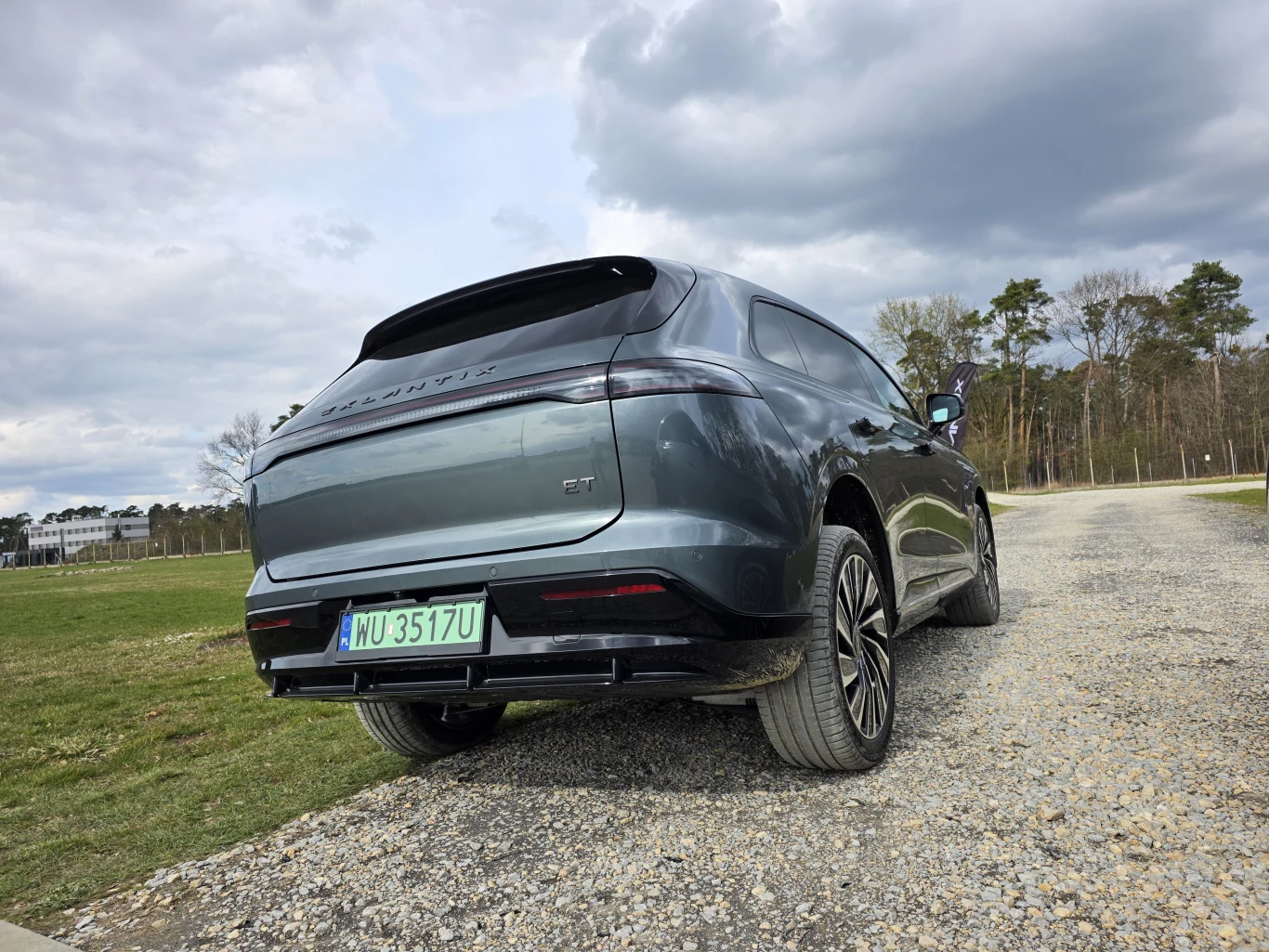 nowoczesny SUV w kolorze metalicznej szarości zaparkowany na szutrowej drodze, z widocznymi dużymi felgami i panoramicznym dachem; wokół samochodu otwarta przestrzeń z trawiastym terenem oraz linią drzew w tle