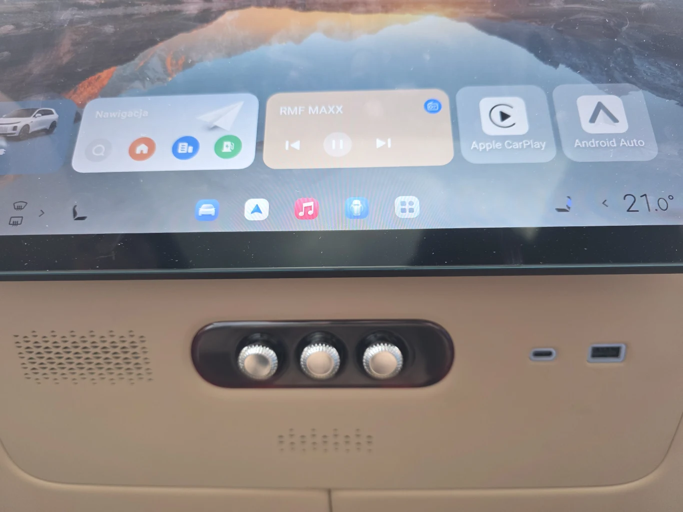 dotykowy panel multimedialny w samochodzie z widocznym ekranem nawigacji, muzyki i opcji Apple CarPlay oraz Android Auto, poniżej trzy fizyczne pokrętła sterowania oraz wentylator