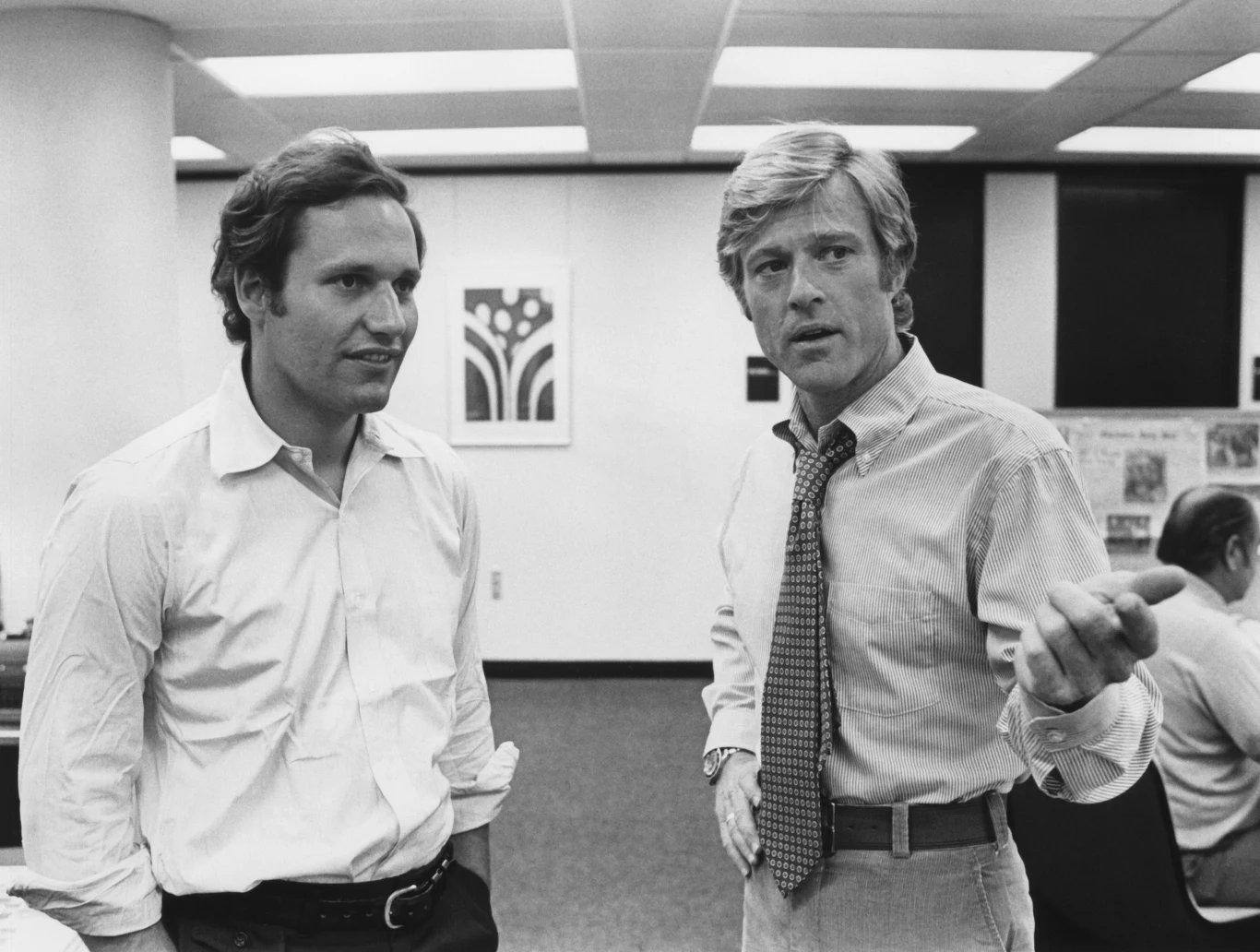 Dwóch mężczyzn w biurowym otoczeniu, ubranych w koszule, jeden z nich gestykuluje, wskazując ręką, podczas gdy drugi patrzy przed siebie; w tle biurka i inni pracownicy.