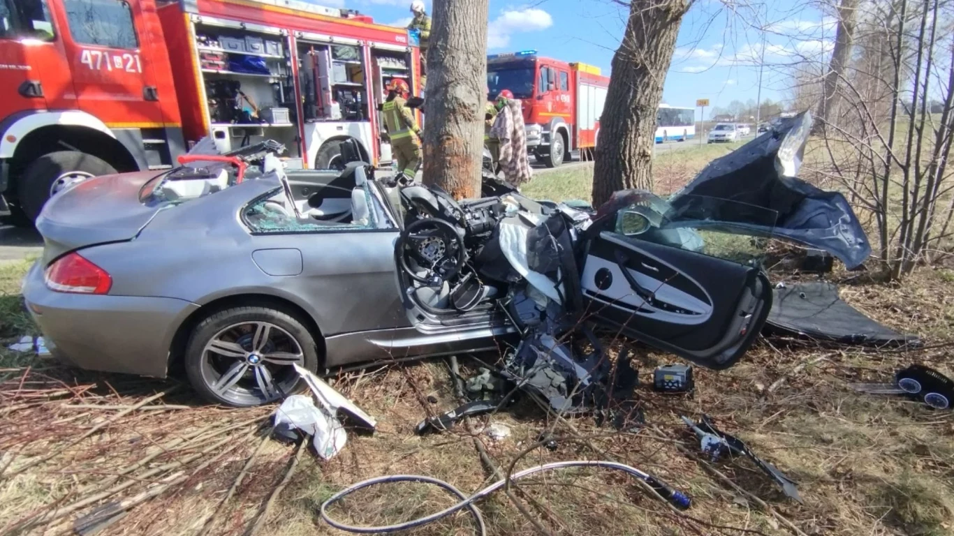 rozbity srebrny samochód osobowy uderzony w bok i prawą stronę w pobliżu drzew, otwarte drzwi i zniszczona karoseria, w tle dwa wozy straży pożarnej oraz strażacy zabezpieczający miejsce wypadku
