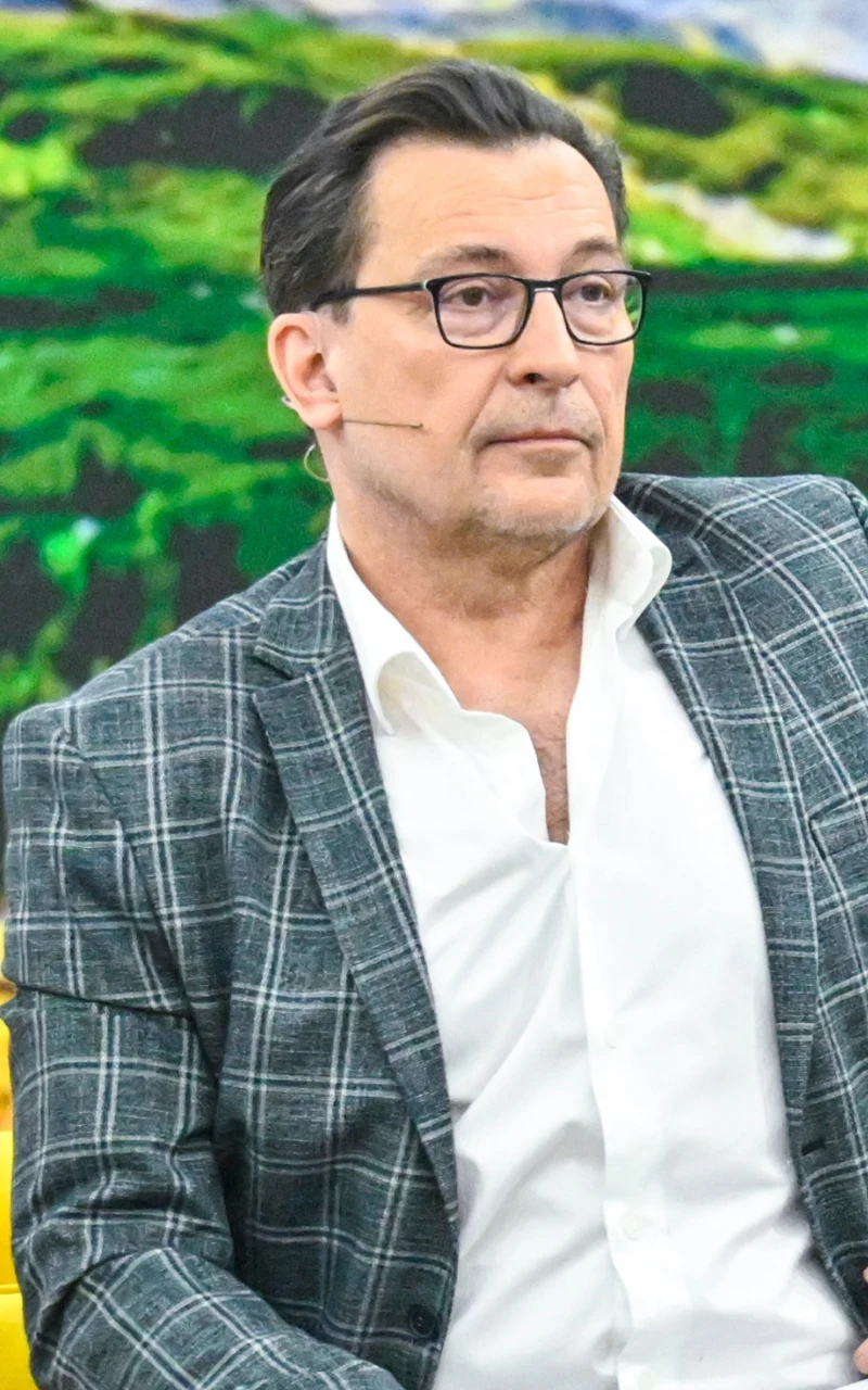 Mężczyzna w okularach i kratowanej marynarce siedzi przed kolorowym, rozmytym tłem zieleni, mając założony mikrofon na twarzy i poważny wyraz twarzy.