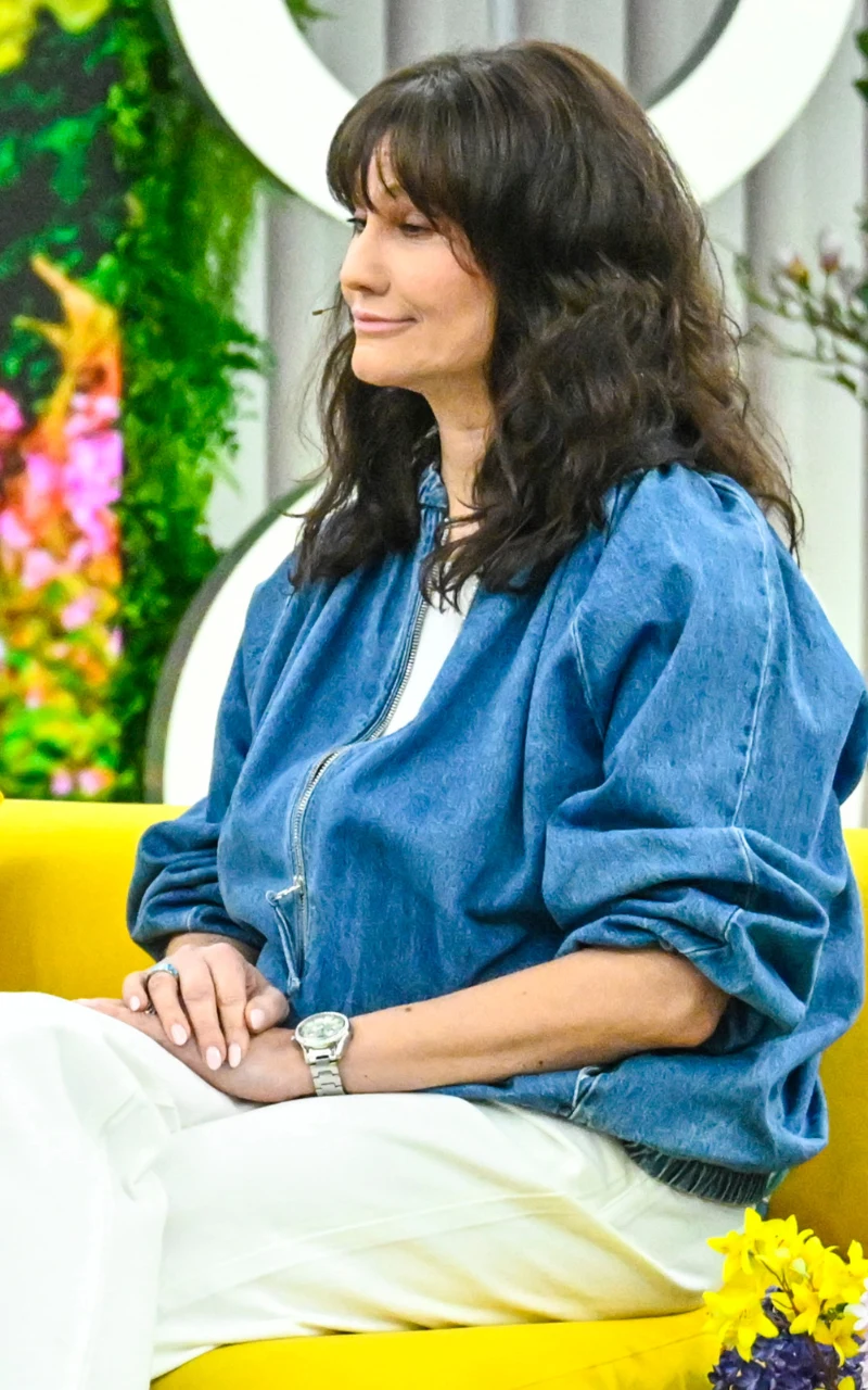 kobieta z długimi, ciemnymi włosami siedząca na żółtej sofie, ubrana w niebieską kurtkę dżinsową i białe spodnie, z widocznym zegarkiem na ręce; w tle kolorowa dekoracja z kwiatów i zielonych roślin