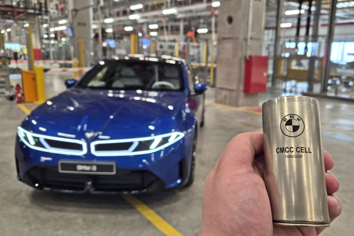 niebieski samochód marki BMW ustawiony w centrum nowoczesnej fabryki, na pierwszym planie w dłoni trzymana cylindryczna bateria z logo BMW i napisem 'CMCC CELL'