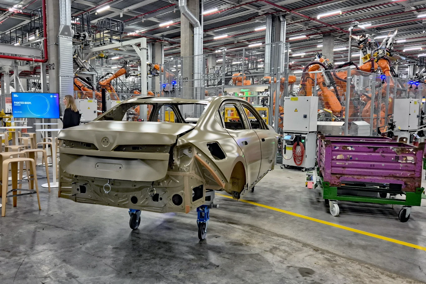 karoseria samochodu osobowego ustawiona na mobilnym stojaku, wokół zaawansowana linia produkcyjna z robotami przemysłowymi wykonującymi operacje montażowe, nowoczesna hala fabryczna pełna maszyn i stanowisk roboczych