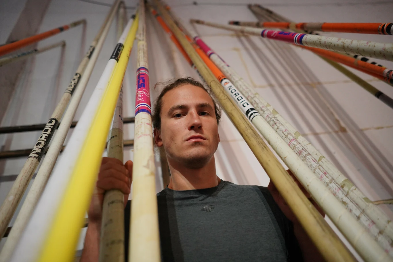 Młody mężczyzna stoi pomiędzy wieloma tyczkami lekkoatletycznymi skierowanymi w górę, trzymając jedną z nich w dłoni; scena skupia się na jego poważnym wyrazie twarzy i różnorodnych kolorach sprzętu sportowego.