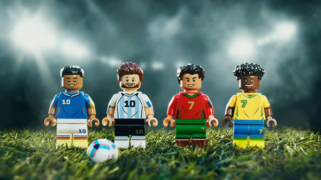 Cztery figurki LEGO piłkarzy ustawione w szeregu na murawie, każda w stroju narodowym reprezentującym inny kraj, z piłką nożną u ich stóp i jasnym oświetleniem stadionowym w tle.