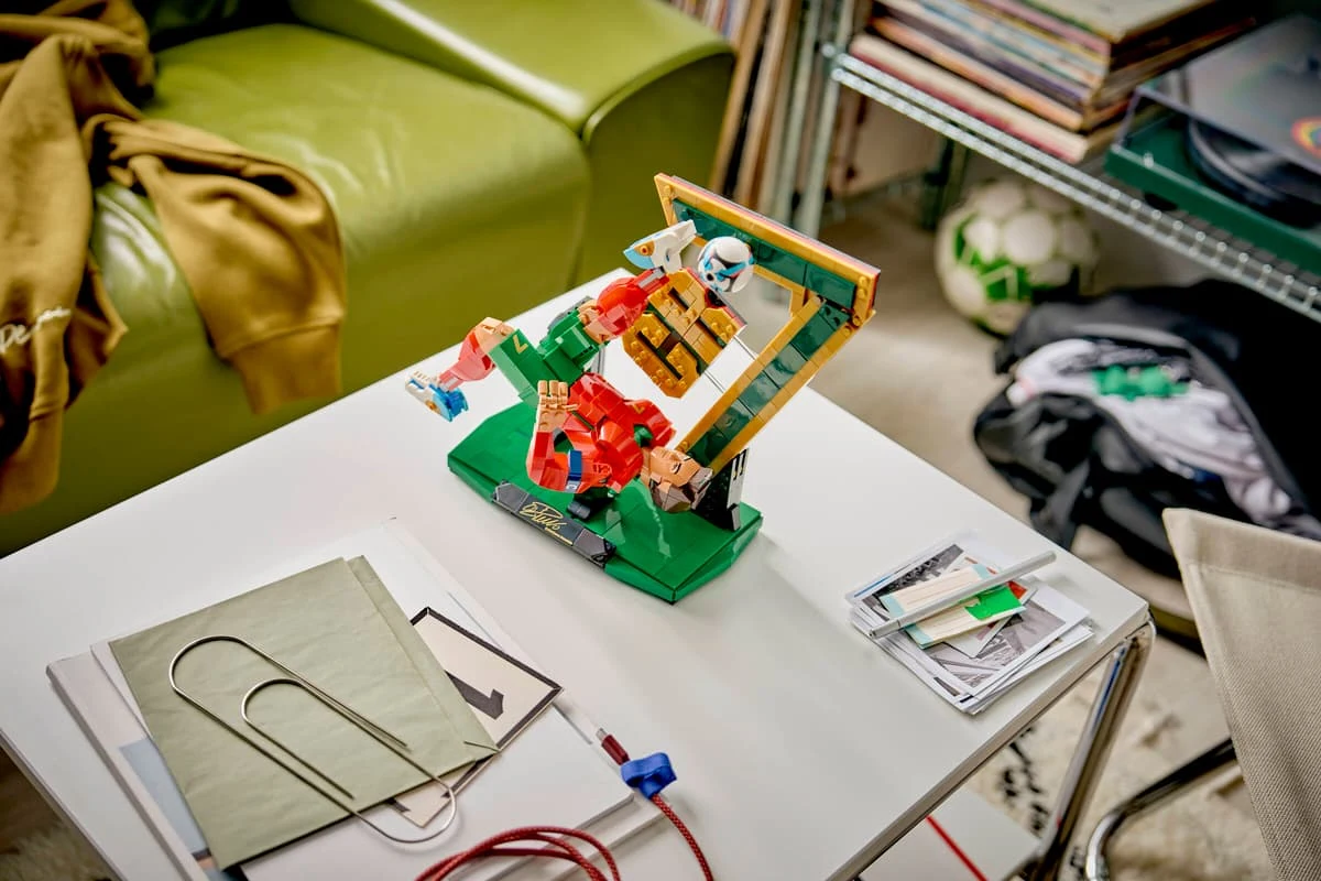 kolorowy zestaw LEGO do gry w piłkarzyki ustawiony na białym stoliku, obok leżą notesy, cienka teczka, spinacz i kilka przewodów, w tle zielona kanapa oraz półka z książkami i winylami