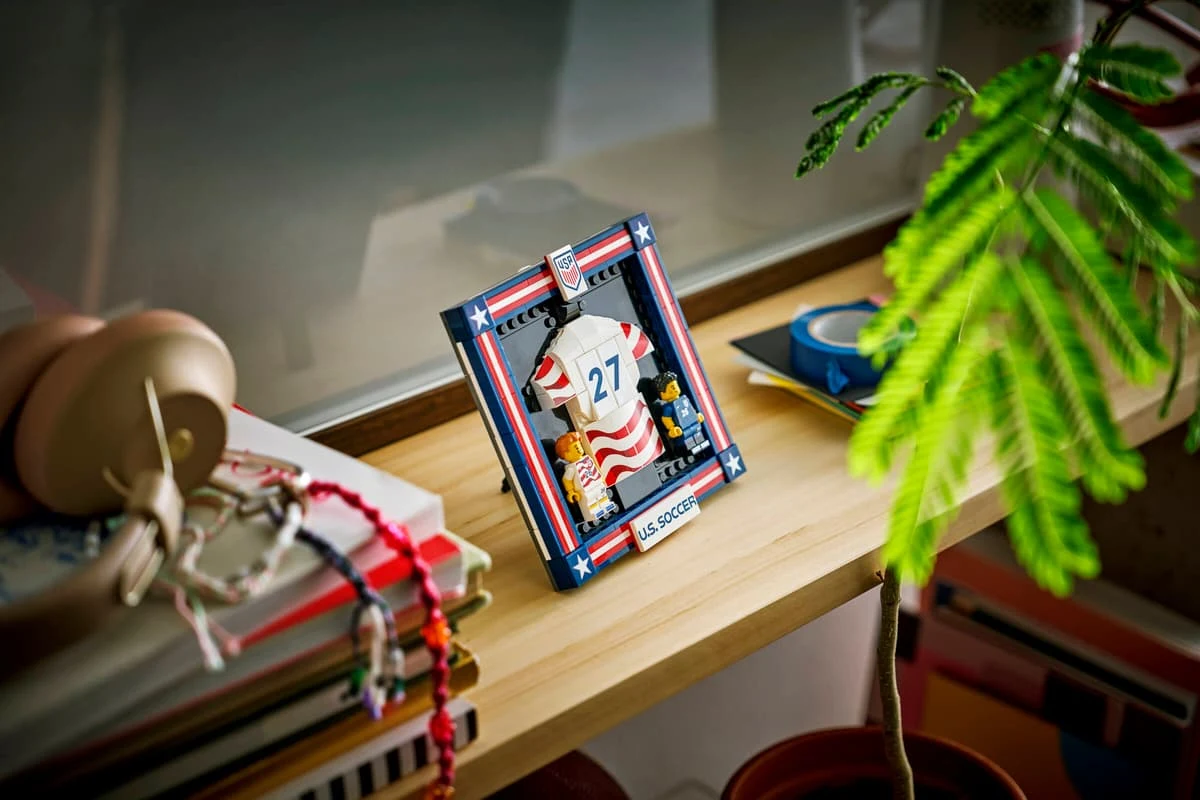 Figurka LEGO w sportowym stroju hokejowym w ramce z numerem 27 ustawiona na drewnianej półce, obok zielonej rośliny i przedmiotów takich jak kolorowe koraliki, stos książek oraz niebieska taśma klejąca.