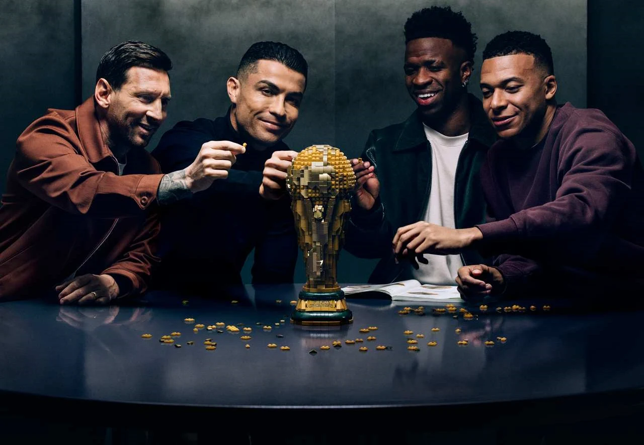 czterech uśmiechniętych mężczyzn siedzi przy stole i wspólnie układa złotą mozaikę w kształcie pucharu świata