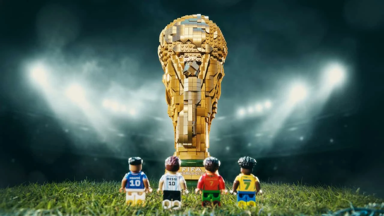 Cztery figurki LEGO w strojach piłkarskich stoją na murawie naprzeciw dużego pucharu FIFA World Cup zbudowanego z klocków, na tle stadionowych reflektorów.