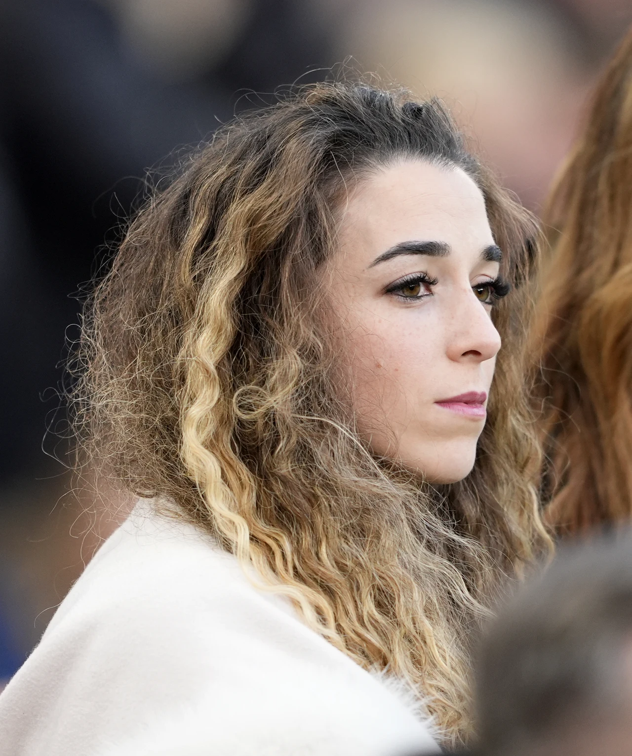 młoda kobieta z długimi, falowanymi włosami w naturalnym odcieniu blond, patrząca w dal z poważnym wyrazem twarzy, ubrana w jasny płaszcz; tło rozmyte, skupienie na twarzy osoby