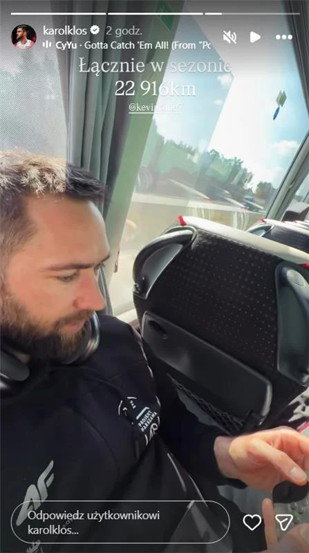 mężczyzna z krótką brodą i słuchawkami na szyi siedzi w autobusie przy oknie, skupiony na ekranie telefonu, za oknem widać rozmyty krajobraz