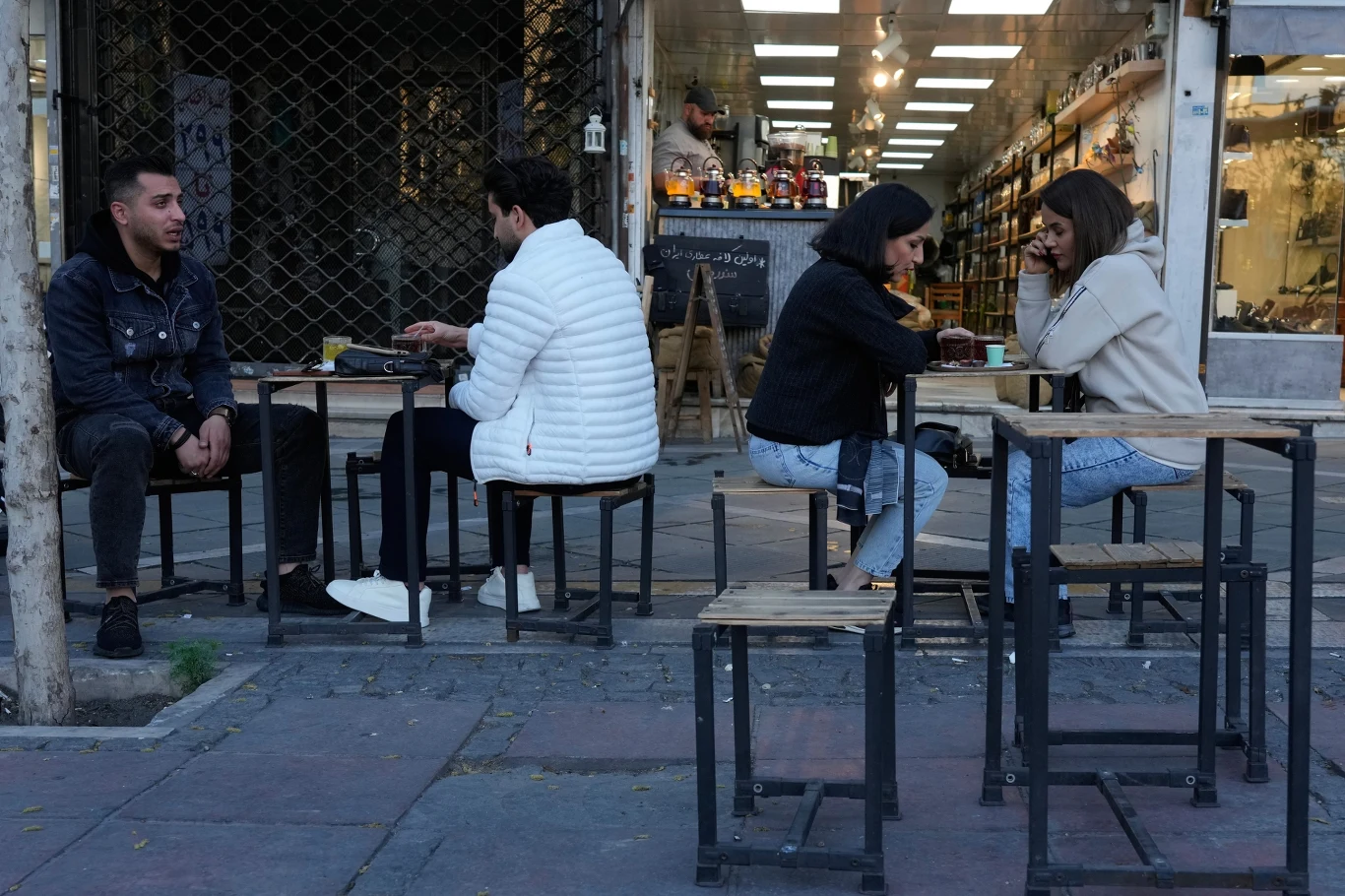 cztery osoby siedzą przy dwóch stolikach na zewnątrz kawiarni, rozmawiają i piją napoje, uliczne otoczenie z otwartym, jasno oświetlonym wnętrzem lokalu w tle