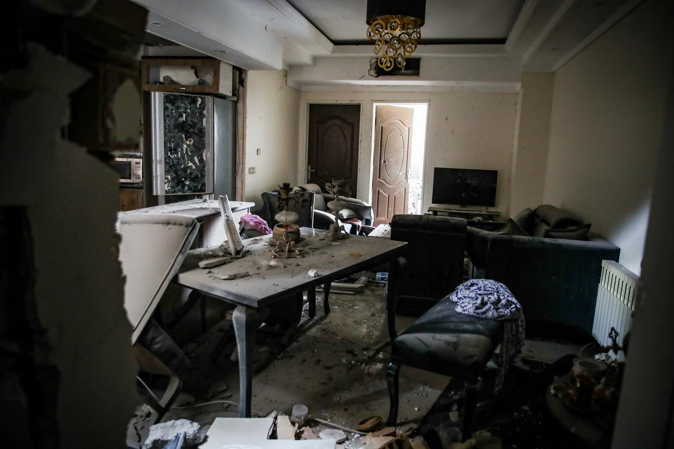 Zniszczony salon z przewróconym stołem, porozrzucanymi meblami i gruzem na podłodze, ślady poważnych uszkodzeń na ścianach i suficie, częściowo zniszczony żyrandol, zdemolowane okno oraz telewizor stojący na szafce.