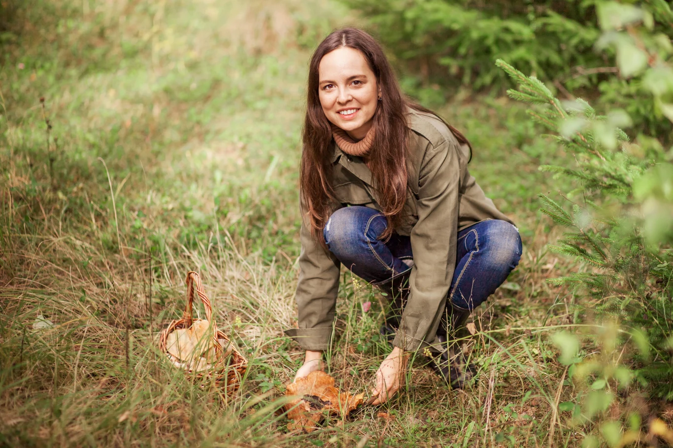 Grzyby pojawiają się w lasach nie tylko jesienią. Sprawdź, jakie grzyby możesz zbierać wiosną i dokąd się udać, by je znaleźć. Kobieta zbiera grzyby w lesie, trzymając dorodny okaz w dłoni. Obok niej, w trawie, wiklinowy kosz. Grzybobranie.