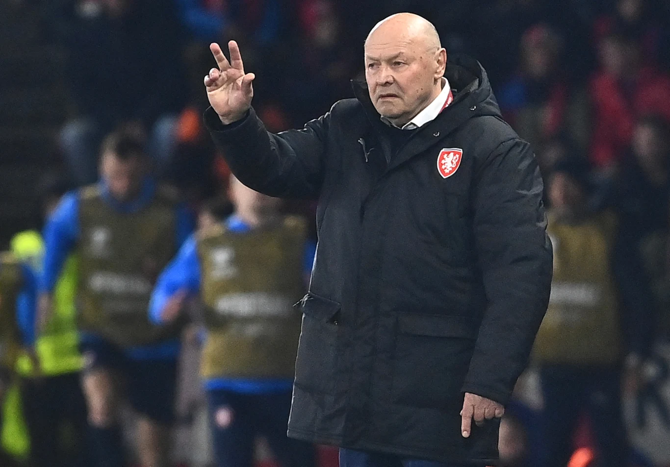 trener drużyny piłkarskiej w czarnej kurtce wykonuje gest ręką, sygnalizując instrukcje podczas meczu, w tle rozmazani piłkarze i sztab szkoleniowy