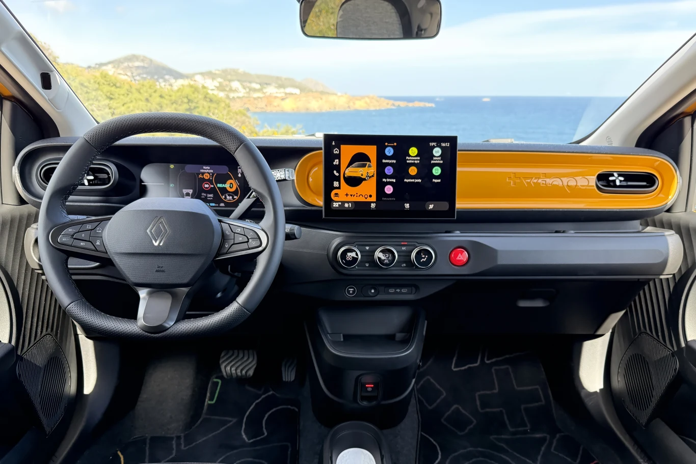 nowoczesny kokpit samochodu marki Renault z cyfrowym zestawem wskaźników, dużym centralnym ekranem dotykowym oraz żółtym wykończeniem deski rozdzielczej, na tle widoczny malowniczy krajobraz nadmorski