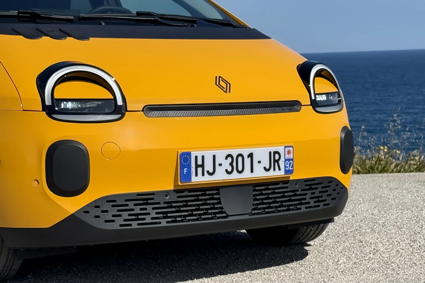 żółty przód nowoczesnego samochodu z charakterystycznymi, prostokątnymi reflektorami LED, minimalistycznym logo na masce oraz francuską tablicą rejestracyjną na tle błękitnego morza