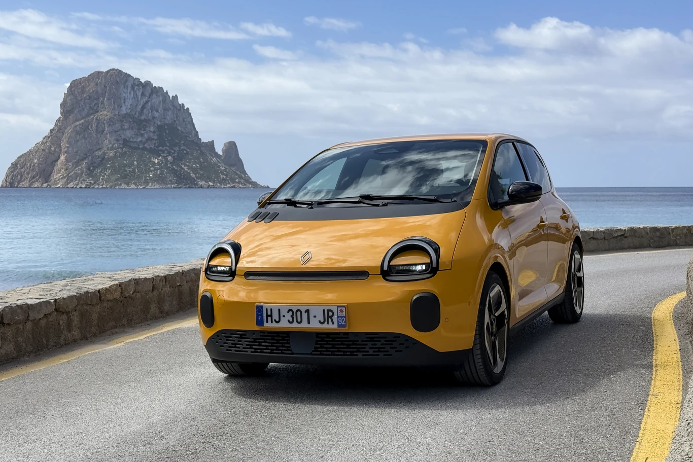 żółty samochód osobowy Renault jadący wzdłuż nadmorskiej drogi z widokiem na skalistą wyspę i spokojne morze w tle