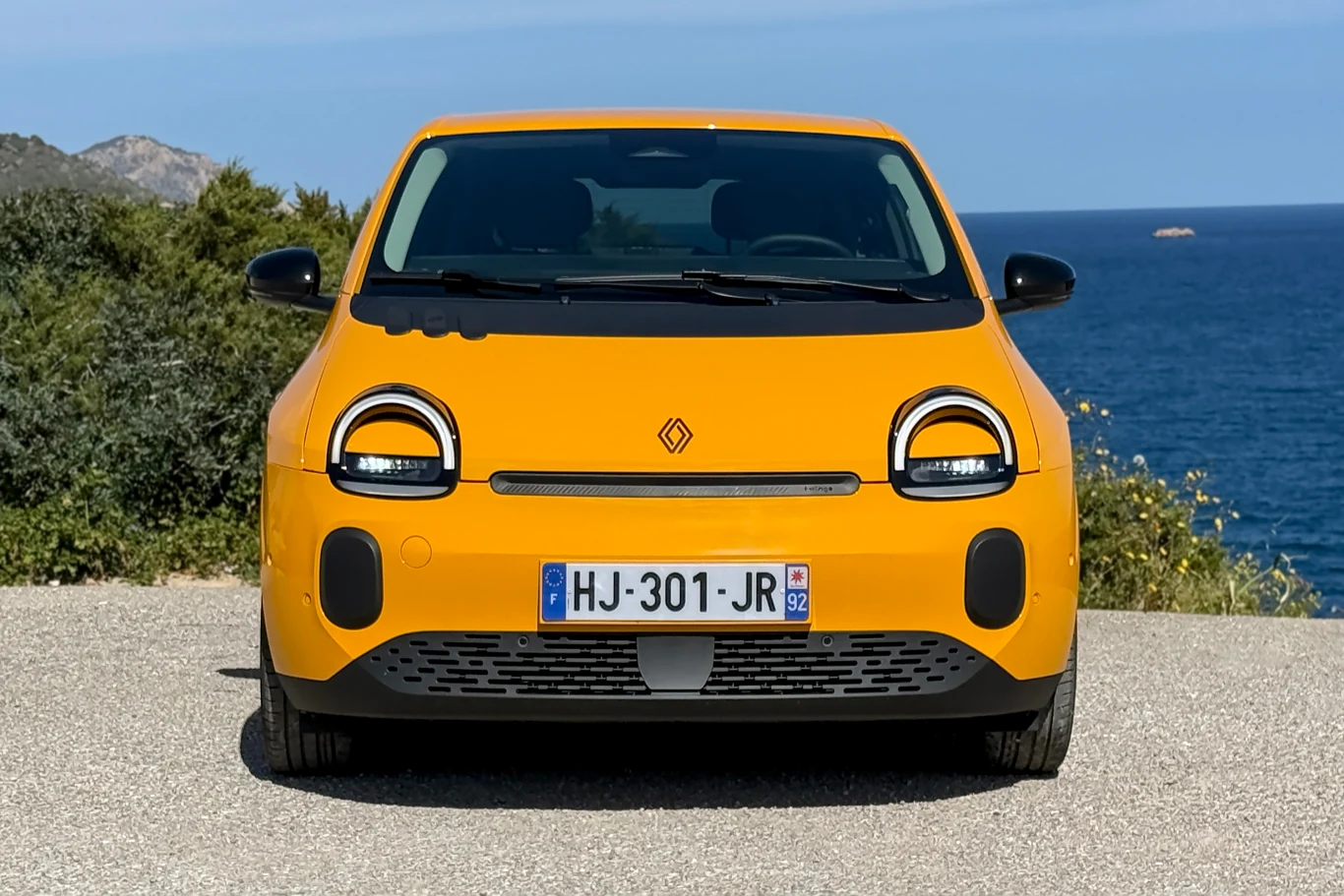 jasnopomarańczowy samochód osobowy o nowoczesnym designie, ustawiony przodem do obserwatora na tle błękitnego morza i zielonych krzewów, z widocznymi charakterystycznymi okrągłymi reflektorami LED, czarnymi detalami i francuską tablicą rejestracyjną